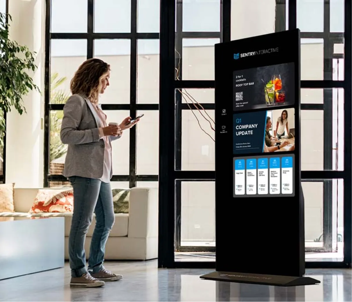 A visitor checks in on a Sentry Interactive kiosk in an office lobby while holding her phone
